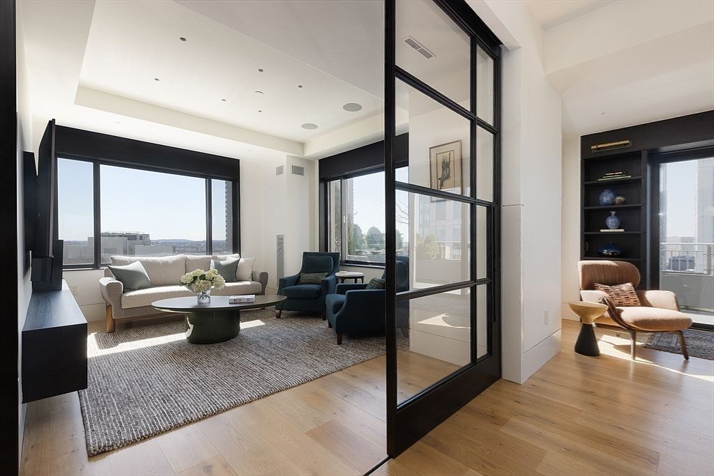 This is an interior shot of a modern living room featuring a neutral-toned sofa, two blue armchairs, and a round coffee table on a gray rug. Large windows provide ample natural light and city views, while a black-framed glass partition separates the living area from another room with a brown armchair and built-in shelving. The hardwood flooring adds warmth to the space, creating an inviting and stylish atmosphere.