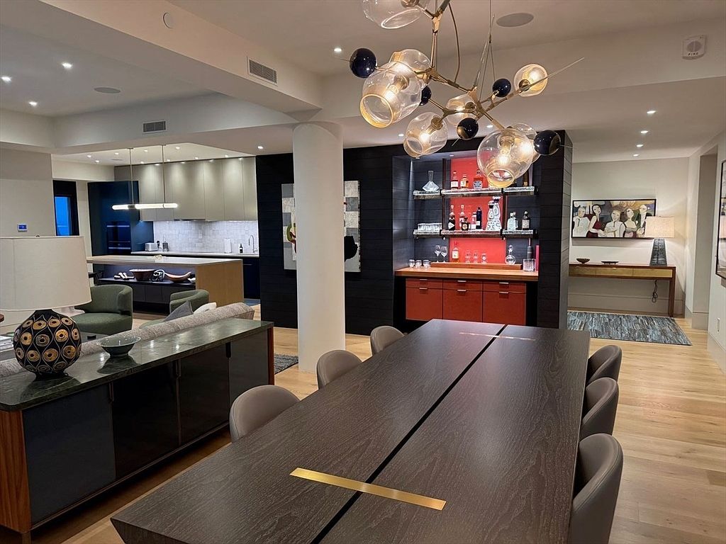 This interior shot showcases a modern dining room with a large, dark wood table featuring a gold accent. The room is well-lit by a contemporary chandelier, and the space flows seamlessly into the living area and kitchen. A built-in bar area with red accents adds a touch of sophistication.