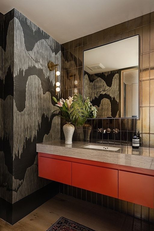 This is a stylish guest bathroom featuring bold wallpaper with a black and silver pattern, complemented by bronze-toned vertical tiles. A floating vanity with red cabinetry and a light-colored countertop adds a pop of color. The mirror reflects the wallpaper, enhancing the depth of the room, and a vase of flowers adds a touch of nature.