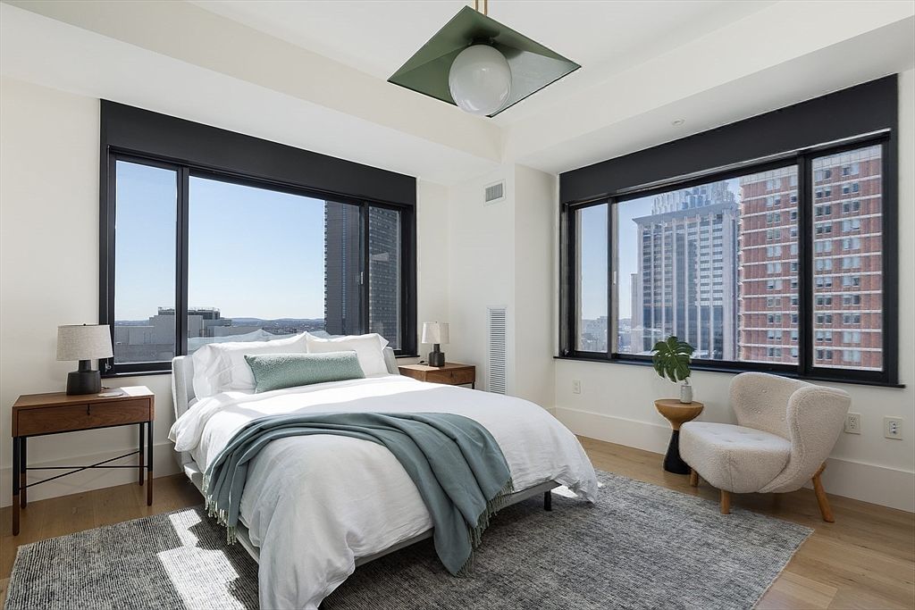This is a bright and modern primary bedroom featuring two large windows with city views. The room is furnished with a bed dressed in white linens and a light blue throw, a wooden nightstand with a lamp, and a cozy armchair. A gray rug anchors the space, and a unique geometric light fixture hangs from the ceiling, creating a stylish and inviting atmosphere.