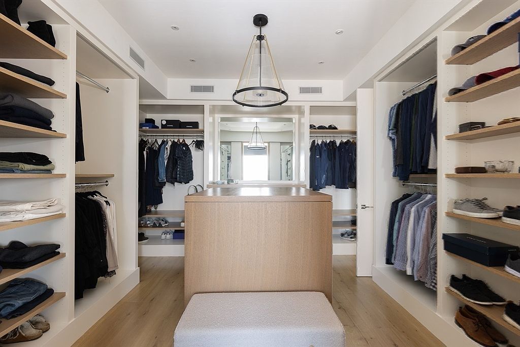 This is a well-organized walk-in closet featuring custom shelving and hanging rods for clothing storage. A central island with a light wood finish provides additional surface space, and a plush ottoman offers a comfortable seating option. The closet is illuminated by a modern pendant light, creating a bright and inviting space.