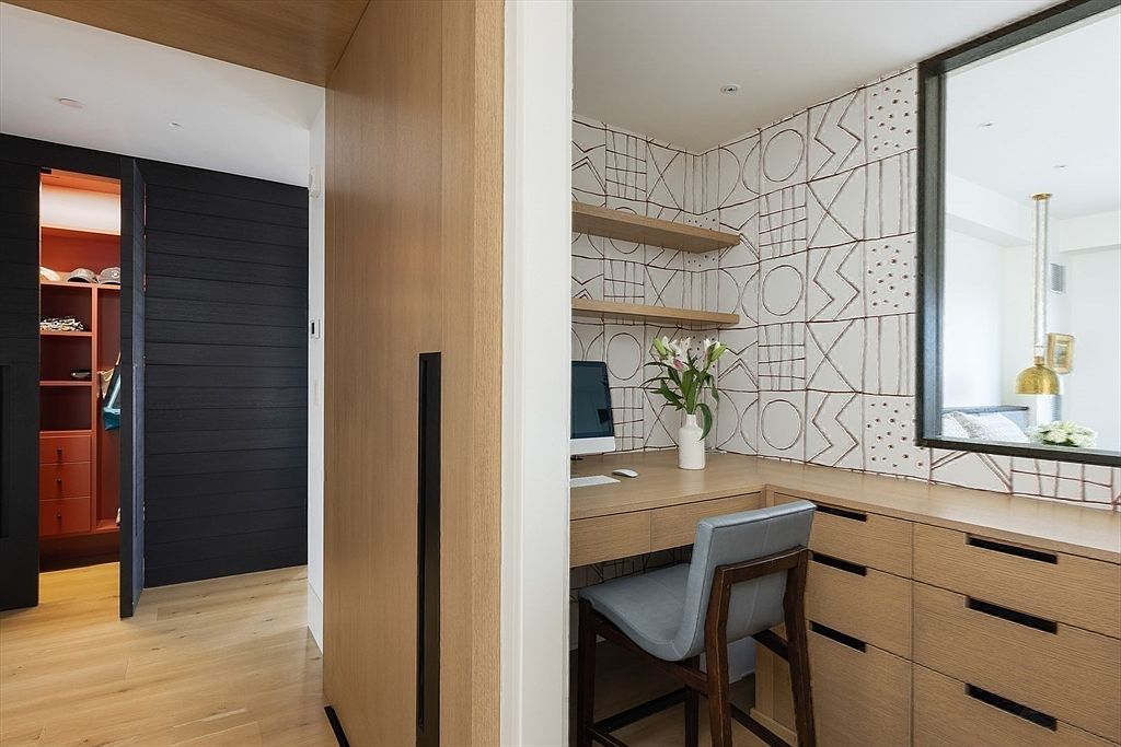 This interior shot showcases a compact yet stylish office space. The area features a built-in desk with drawers, open shelving, and a unique patterned tile wall. A comfortable chair and a glimpse of a closet through an adjacent doorway add to the functionality and appeal of this workspace.