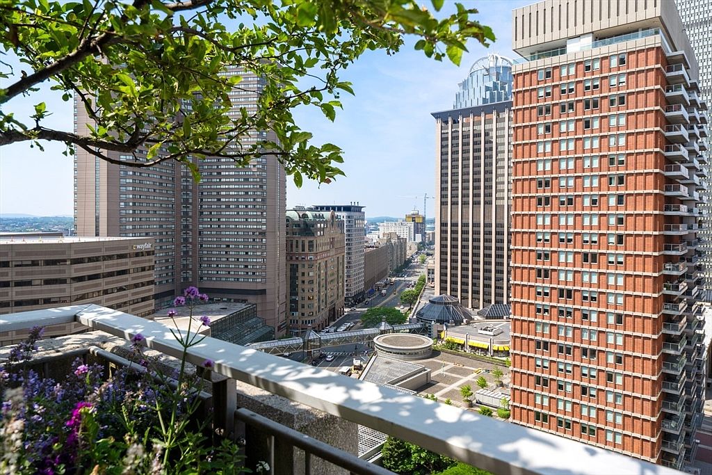 This image showcases a stunning cityscape view from a high-rise balcony. The balcony features a stone railing with metal accents and a planter box filled with vibrant purple flowers. The view includes several modern skyscrapers, a bustling street below, and a clear blue sky, creating an impressive urban panorama.