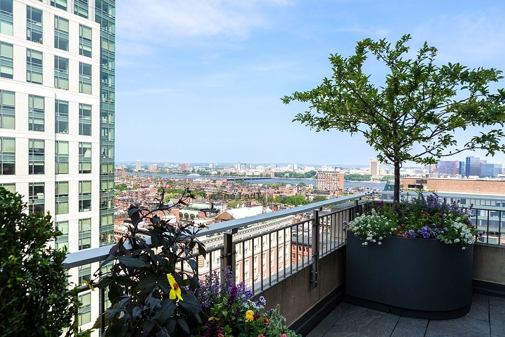 This image showcases a luxurious balcony with stunning city views. The balcony features a modern railing, elegant planters with vibrant flowers and a small tree, and a sleek, dark-toned flooring. A tall building is visible on the left, adding to the urban landscape.