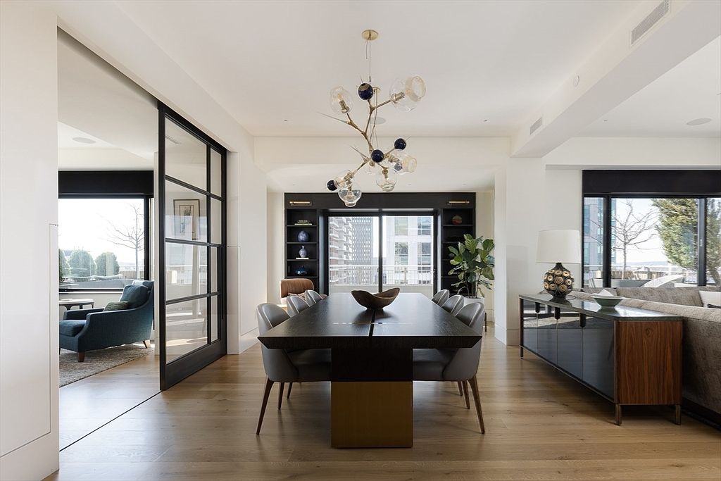 This is an interior shot of a modern dining room. The room features a long, dark wood dining table with gray chairs, illuminated by a unique, sculptural chandelier. Large windows offer city views, and the open layout connects to other living spaces, enhancing the sense of spaciousness and luxury.
