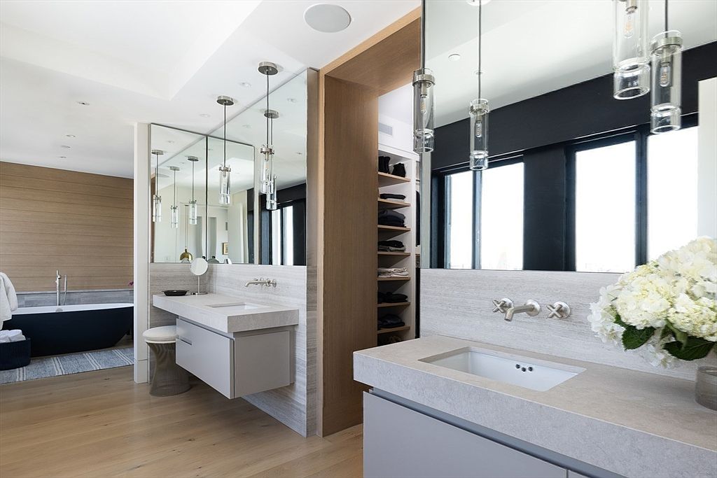 This is a modern primary bathroom featuring dual vanities with light gray cabinets and light stone countertops. Large mirrors reflect pendant lighting and natural light from a nearby window, enhancing the spacious feel. A separate area with a freestanding tub is visible in the background, adding to the luxurious ambiance.