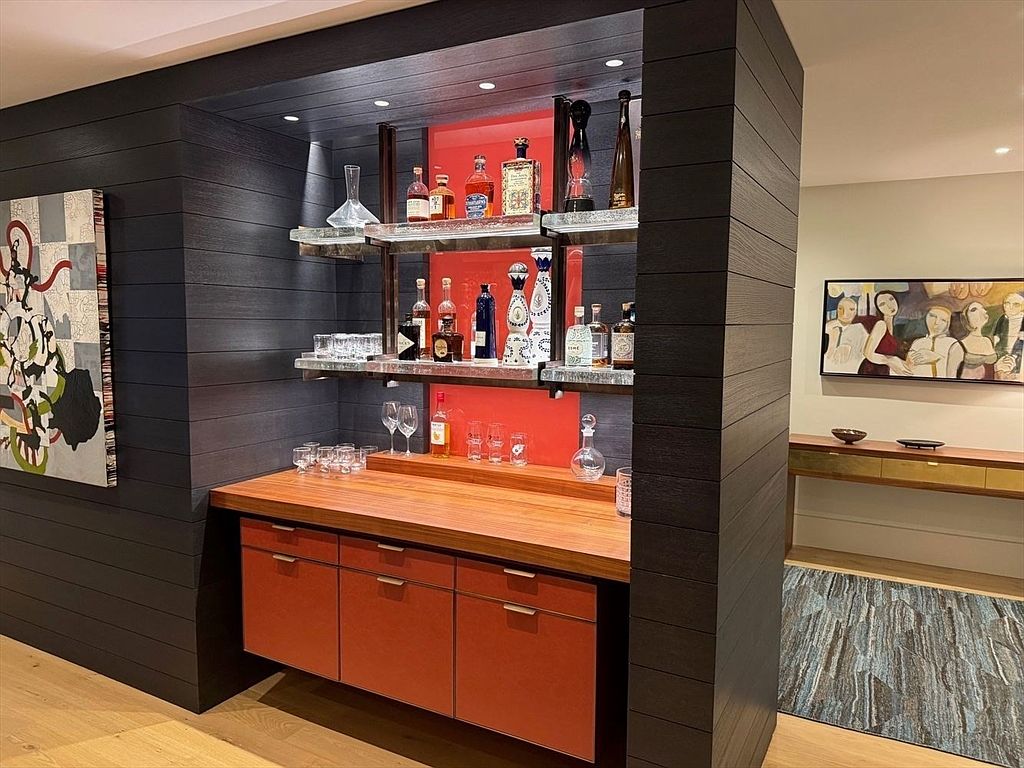 This image showcases a stylish home bar area integrated into a living space. The bar features dark wood paneling, glass shelves displaying various bottles and glassware, and a warm-toned countertop with orange cabinetry below. Artwork on the adjacent wall adds a touch of sophistication.