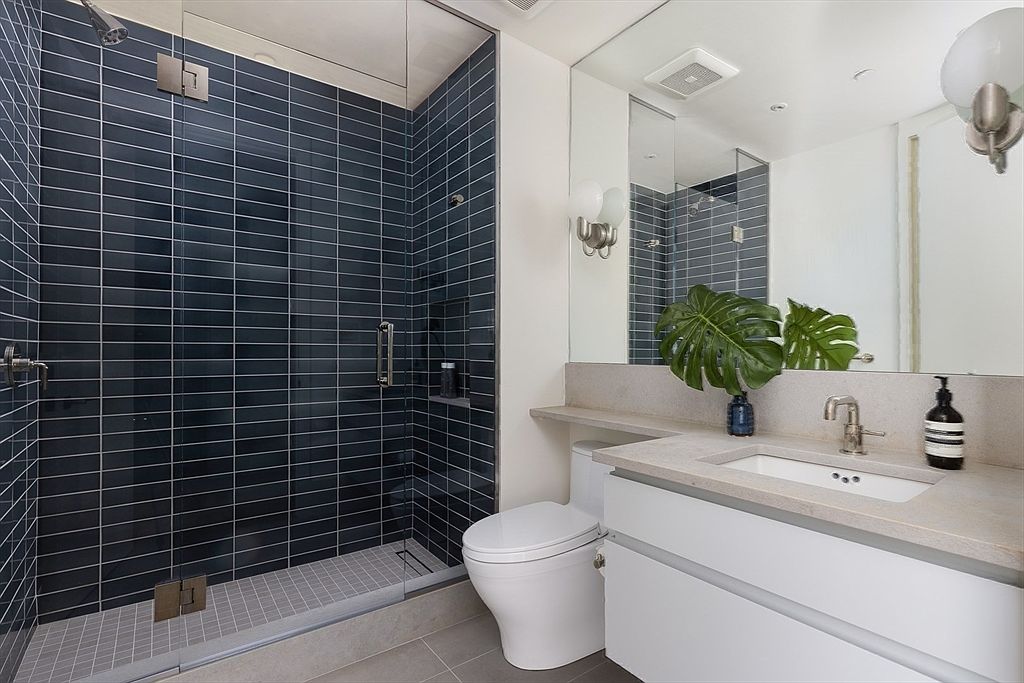This is a modern bathroom featuring a walk-in shower with dark blue subway tiles and a glass enclosure. The vanity has a light-colored countertop and white cabinets, complemented by a large mirror and contemporary lighting fixtures. A white toilet is positioned next to the vanity, and the overall design is clean and minimalist.