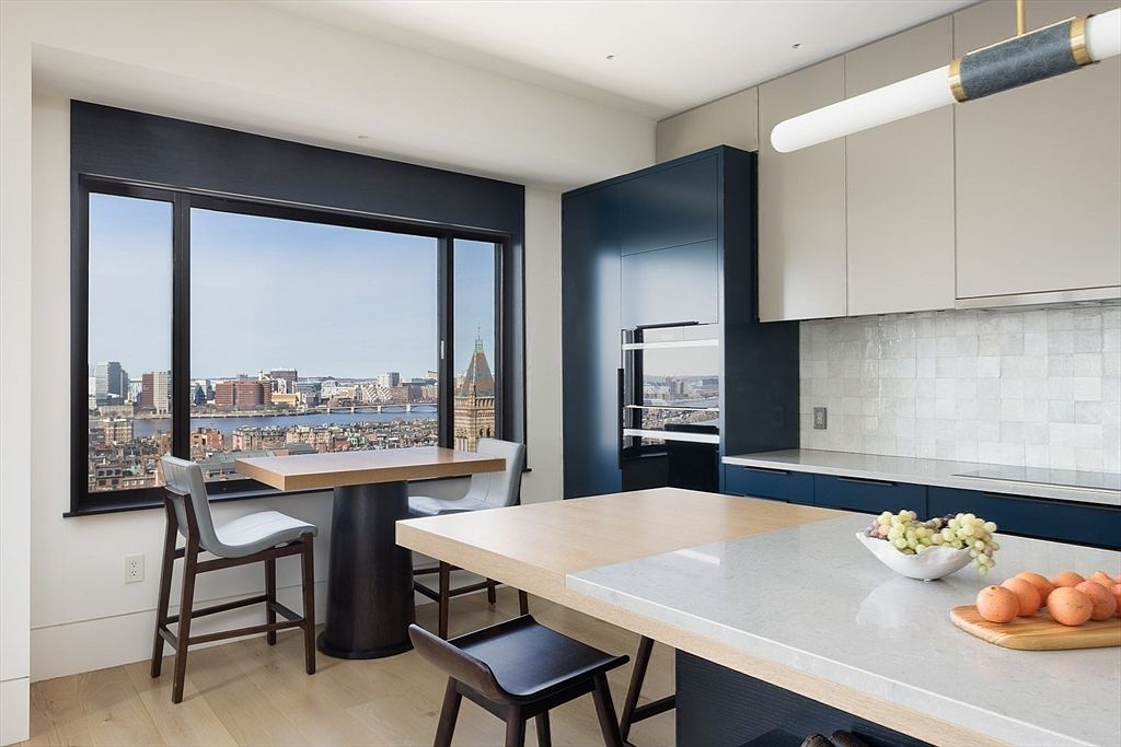 This is a modern kitchen with a stunning city view from a large window. The kitchen features a combination of blue and white cabinetry, a kitchen island with a light countertop, and a dining table near the window. The overall design is sleek and contemporary, emphasizing natural light and urban living.