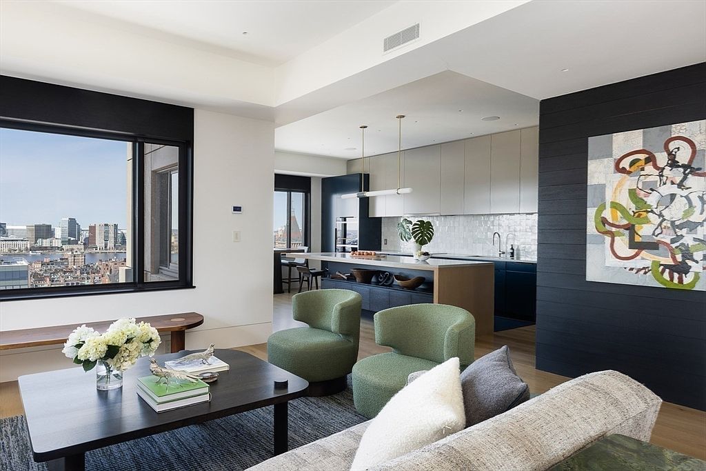 This is an interior shot of a modern living room that seamlessly blends into a kitchen area. The living room features a dark wood coffee table, green armchairs, and a comfortable sofa with decorative pillows. The kitchen boasts sleek cabinetry, a large island with bar seating, and modern pendant lighting, creating an open and inviting space with a view of the city skyline.