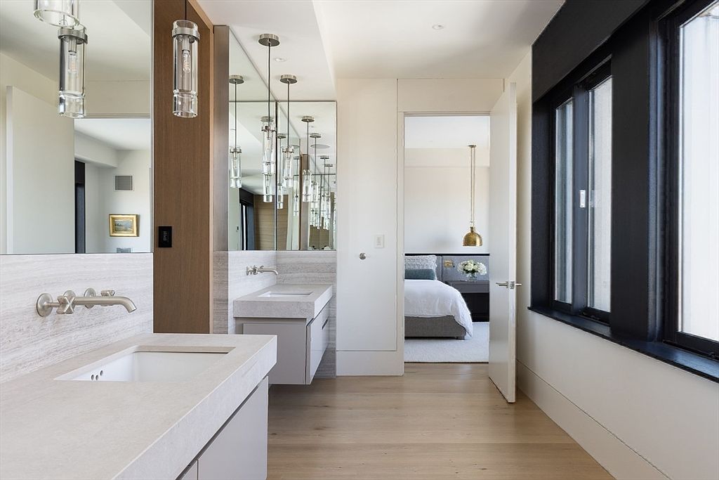 This is a modern primary bathroom featuring dual vanities with light-colored countertops and cabinetry. Large mirrors are illuminated by pendant lighting, and the space flows into an adjacent bedroom, visible through an open doorway. The flooring is light hardwood, and a large window with black trim provides natural light.