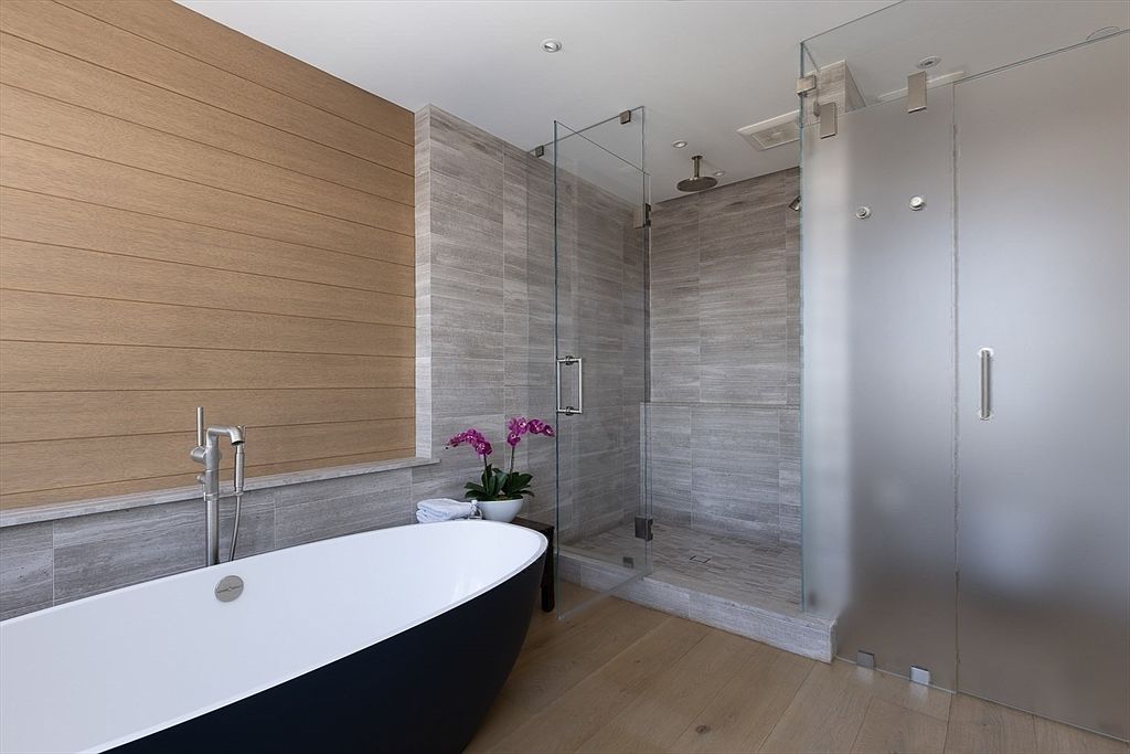 This is a modern primary bathroom featuring a freestanding black and white bathtub with a sleek faucet. The bathroom has a glass-enclosed shower with gray tiled walls and a frosted glass door. The walls are a combination of wood paneling and gray tile, and the floor is light wood, creating a spa-like atmosphere.