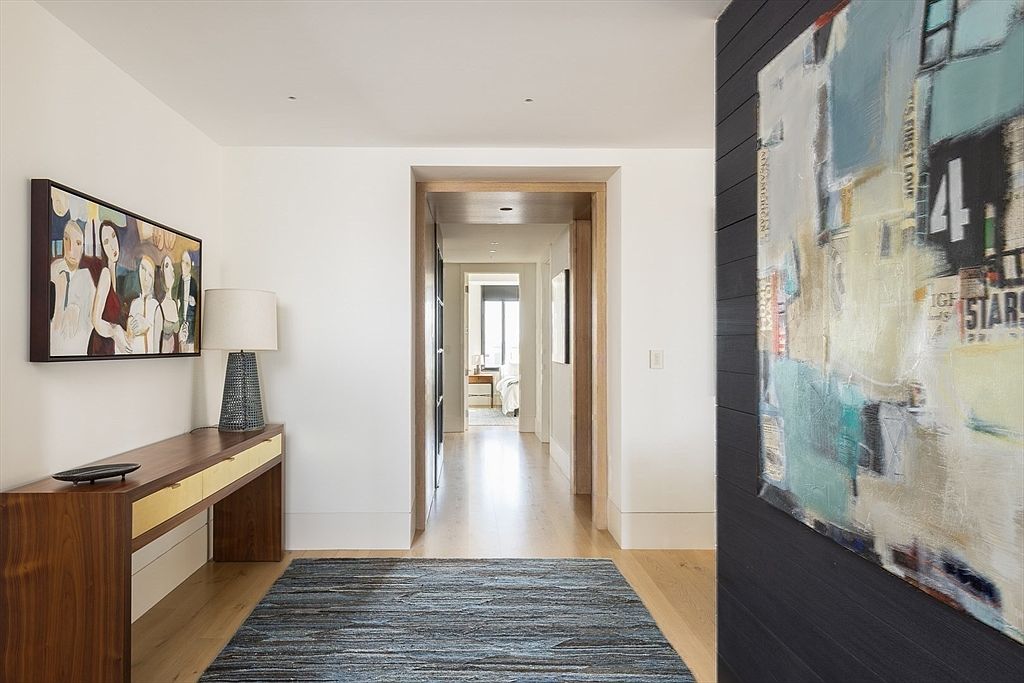 This interior shot showcases a well-lit hallway with hardwood floors and white walls, creating a clean and modern aesthetic. A console table with a lamp and artwork adorns one side, while a large abstract painting adds visual interest to the other. The hallway leads to another room, framed by a wooden doorway, suggesting a seamless flow within the home.
