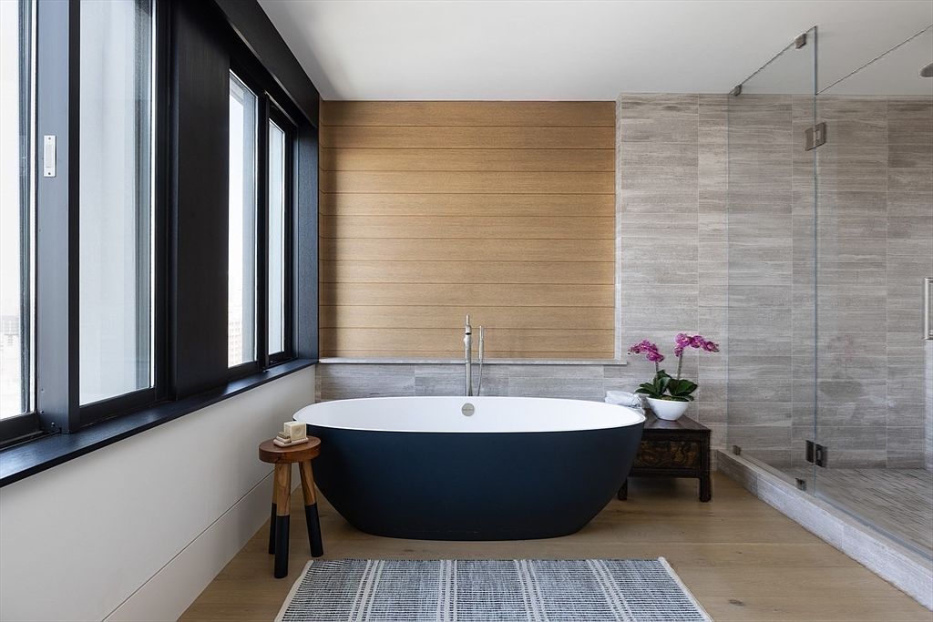 This is a luxurious primary bathroom featuring a freestanding black and white bathtub as the focal point, complemented by a wooden stool and a striped rug. Large windows with black frames provide natural light, while a wood-paneled wall and tiled shower area add texture and depth. The overall impression is modern, serene, and spa-like.