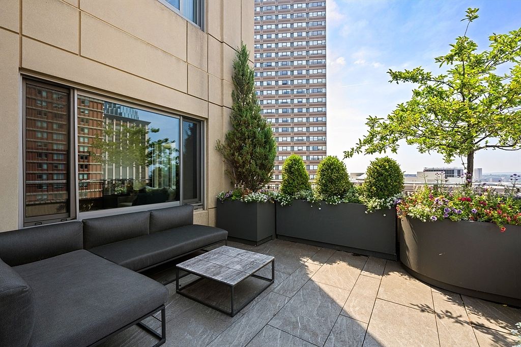This image showcases a stylish balcony or patio area, featuring a modern outdoor sofa and a sleek coffee table. The space is enhanced by large planters filled with greenery and flowers, creating a serene and inviting atmosphere. The backdrop includes a view of a neighboring building, adding an urban touch to the outdoor living space.