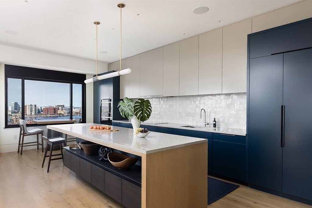 This is a modern kitchen featuring two-tone cabinetry in navy and white, with a large island topped with a light-colored countertop. The island has a wooden base and dark storage cabinets, and is adorned with decorative wooden bowls. A large window offers a city view, and modern pendant lighting hangs above the island.