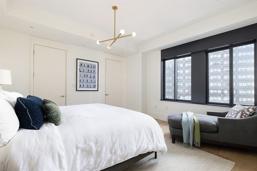 This is a bright and airy primary bedroom featuring a large bed with white linens and decorative pillows. The room has a modern chandelier, a framed art piece, and large windows with dark frames that offer city views. A chaise lounge sits near the window, adding a touch of luxury and comfort to the space.