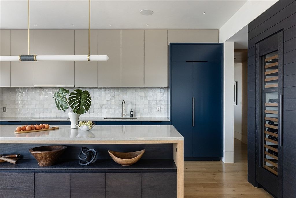 This is a modern kitchen featuring two-tone cabinetry in beige and navy blue, complemented by a white countertop island. The kitchen is illuminated by a linear pendant light, and the backsplash is a textured tile. A wine fridge is integrated into the dark wood paneling on the right, adding a touch of luxury.
