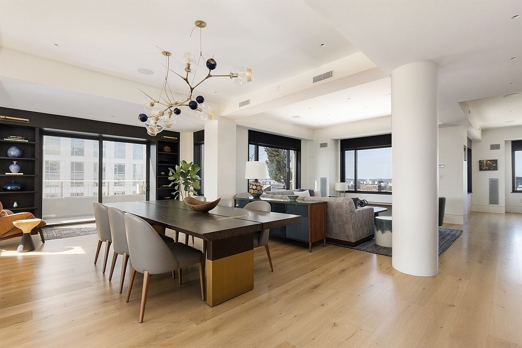 This is a spacious and modern living and dining area featuring hardwood floors, large windows with black frames, and a contemporary chandelier. The dining area includes a dark wood table with light gray chairs, while the living area has a comfortable gray sofa and stylish accent furniture. A large white column adds architectural interest to the open-concept space.