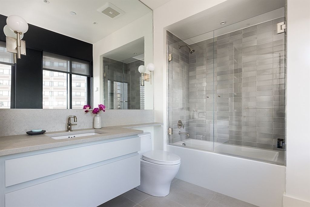 This is a modern primary bathroom featuring a sleek vanity with a light-colored countertop and white drawers, a toilet, and a bathtub enclosed by a glass shower screen with gray subway tile. The room is well-lit, and the overall design is clean and contemporary, creating a spa-like atmosphere. A large mirror reflects the window and the city view outside.