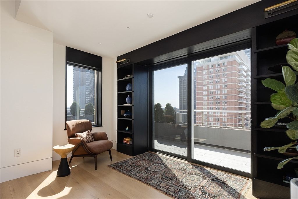 This is a bright and stylish living room featuring a modern aesthetic. The room includes a comfortable armchair, a unique side table, and a patterned rug that adds warmth to the space. Large sliding glass doors lead to a balcony, offering city views and natural light, complemented by built-in shelving units on either side.