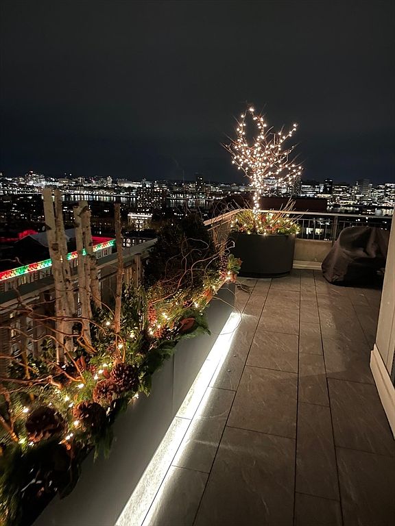 This image showcases a beautifully decorated balcony at night, featuring illuminated planters and a small tree adorned with lights. The balcony's design incorporates modern gray tiling and a sleek, minimalist aesthetic, while the city skyline provides a stunning backdrop. The overall impression is one of sophisticated urban living and outdoor enjoyment.