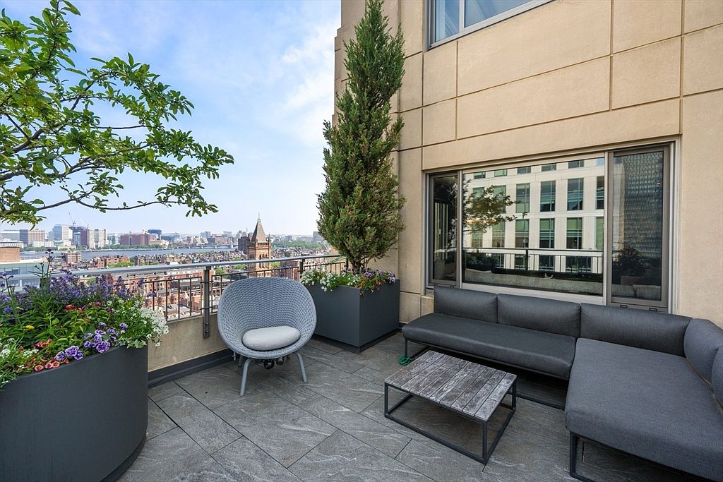 This image showcases a stylish balcony with a cityscape view. The balcony features modern outdoor furniture, including a gray sofa, a round woven chair, and a wooden coffee table. Potted plants and a small tree add greenery to the space, creating a relaxing and inviting atmosphere.