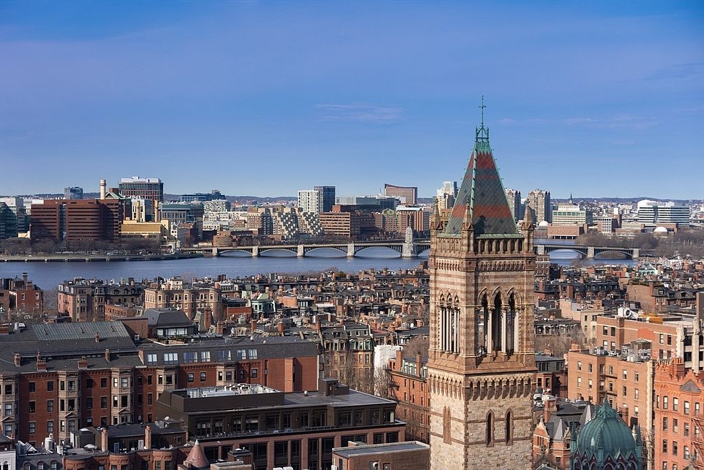 This aerial view showcases a cityscape with a prominent church tower in the foreground. The city features a mix of brick buildings and modern structures, with a river and bridges visible in the midground. The clear blue sky provides a bright backdrop, highlighting the urban landscape and architectural details.