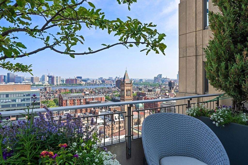 This image showcases a luxurious balcony with stunning city views. The balcony features a comfortable wicker chair, lush flower planters, and a wrought iron railing. The cityscape includes prominent buildings and a clear blue sky, creating an inviting and upscale outdoor living space.