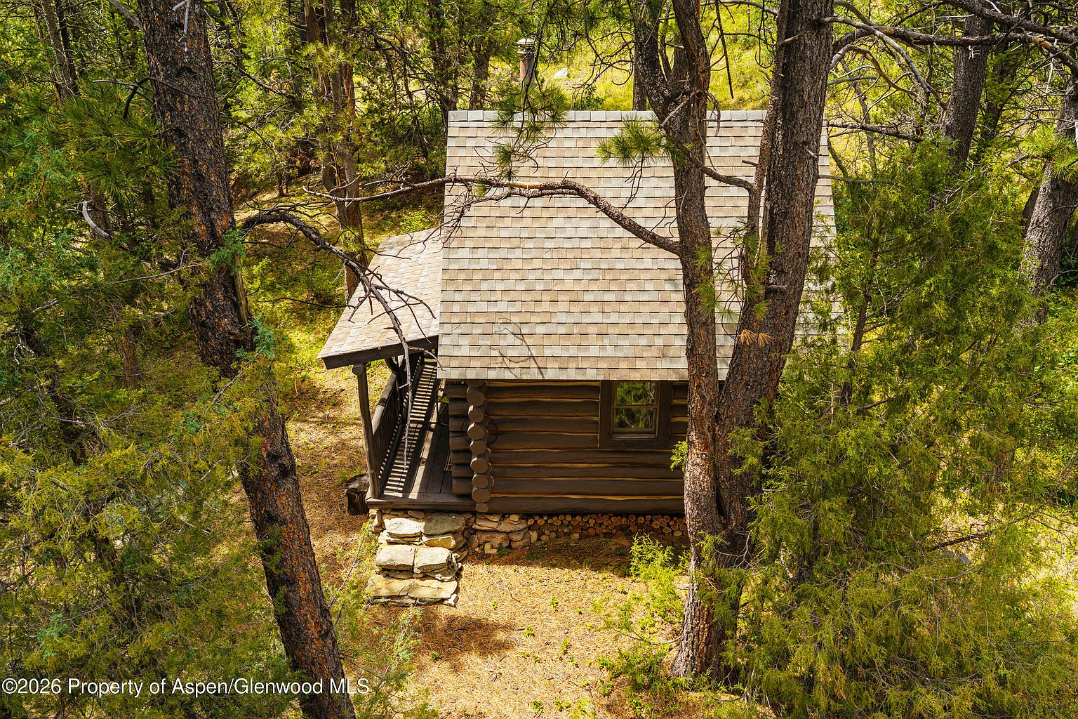 This high-angle aerial shot captures a rustic log cabin nestled deep within a dense, wooded forest. The structure features a shingled roof, a small covered porch with wooden railings, and a stone foundation base. The surrounding trees provide a secluded, natural setting, emphasizing the cabin's quiet and private atmosphere.