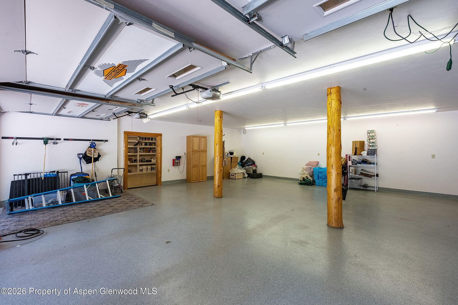 This spacious, multi-car garage features a durable, speckled epoxy floor and a high ceiling with exposed tracks and a Harley-Davidson emblem on the door panel. Two prominent, rustic log support columns anchor the center of the room, while a built-in wooden storage cabinet and a recessed shelving unit provide organized utility space. The area is brightly lit by long, overhead fluorescent light fixtures, creating a clean and functional environment for vehicle storage or workshop activities.