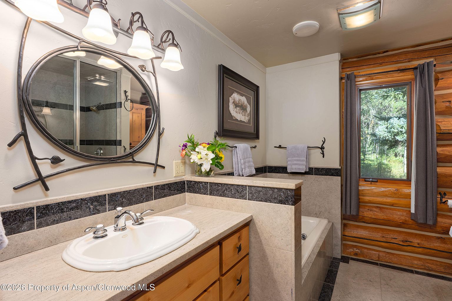 This rustic-style bathroom features a vanity with a light wood cabinet and a neutral stone countertop, complemented by a decorative oval mirror with a branch-like frame. A half-wall separates the vanity area from a soaking tub, which is positioned next to a window framed by log walls and grey curtains. The space is accented with dark, patterned tile trim and warm lighting, creating a cozy and natural atmosphere.