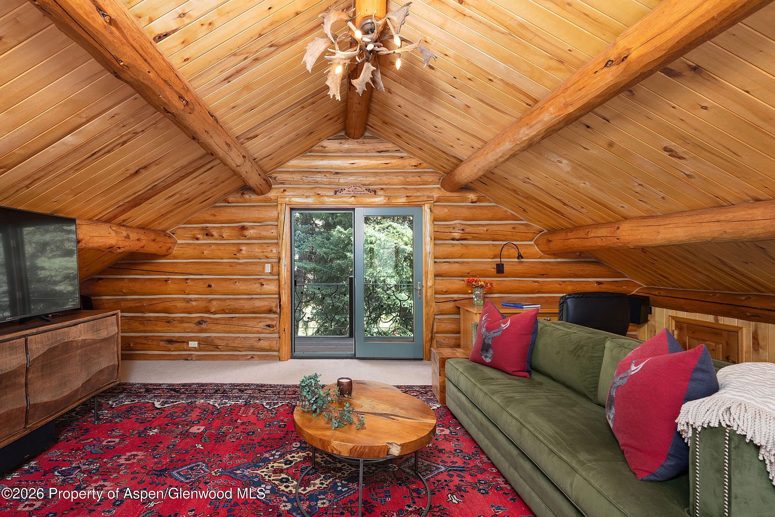 This cozy, rustic living room is situated in an attic space with vaulted ceilings featuring exposed log beams and wood-paneled walls. The room is furnished with a plush green sofa adorned with decorative pillows, a circular wood coffee table, and a television console, all set atop a large, ornate red patterned rug. A glass door leads out to a balcony, allowing natural light to fill the space, while an antler-style chandelier adds a distinct mountain-lodge aesthetic.