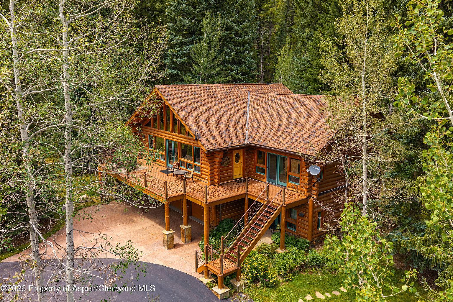 This elevated aerial perspective captures a rustic log cabin nestled within a dense, lush forest. The home features a prominent wrap-around wooden deck with decorative metal railings, a paved driveway area underneath, and a set of stairs leading to the main level. The structure is characterized by its natural log construction, a multi-pitched roof with brown shingles, and large windows that reflect the surrounding greenery.