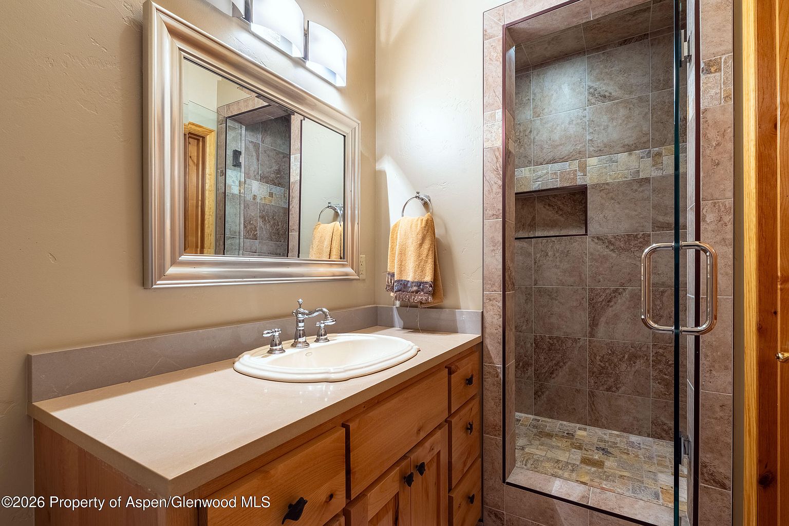 This bathroom features a light wood vanity with a solid-surface countertop and an integrated sink, complemented by a large, framed mirror and a modern three-light fixture. To the right, a glass-enclosed shower is finished with neutral-toned wall tiles and a built-in recessed niche. The space is warm and functional, showcasing a clean, traditional design aesthetic.