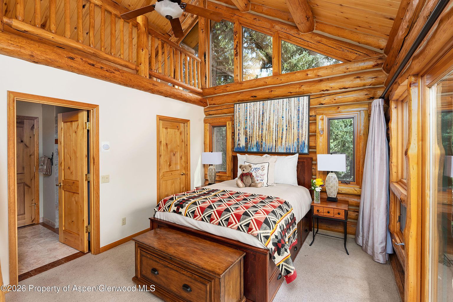 This cozy bedroom features rustic log walls and a vaulted ceiling with exposed timber beams, creating a warm cabin atmosphere. The room is centered around a bed adorned with a patterned quilt and a teddy bear, flanked by two matching bedside lamps and a wooden storage chest at the foot. Large windows and a lofted balcony area above add architectural interest and allow natural light to fill the space.