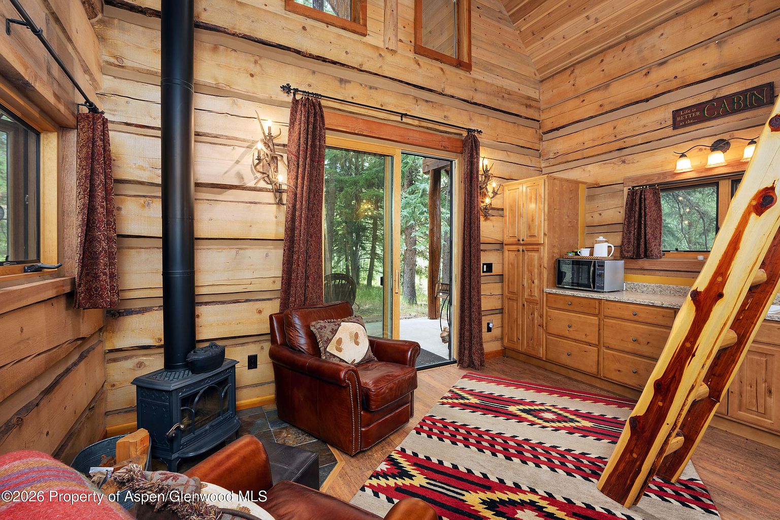 This cozy cabin living area features rustic log walls and a warm, inviting atmosphere centered around a black wood-burning stove. A leather armchair sits near a sliding glass door that opens to a wooded view, while a patterned area rug adds texture to the space. To the right, a small kitchenette area with wooden cabinetry and a microwave provides functional convenience, complemented by a decorative sign above the counter.