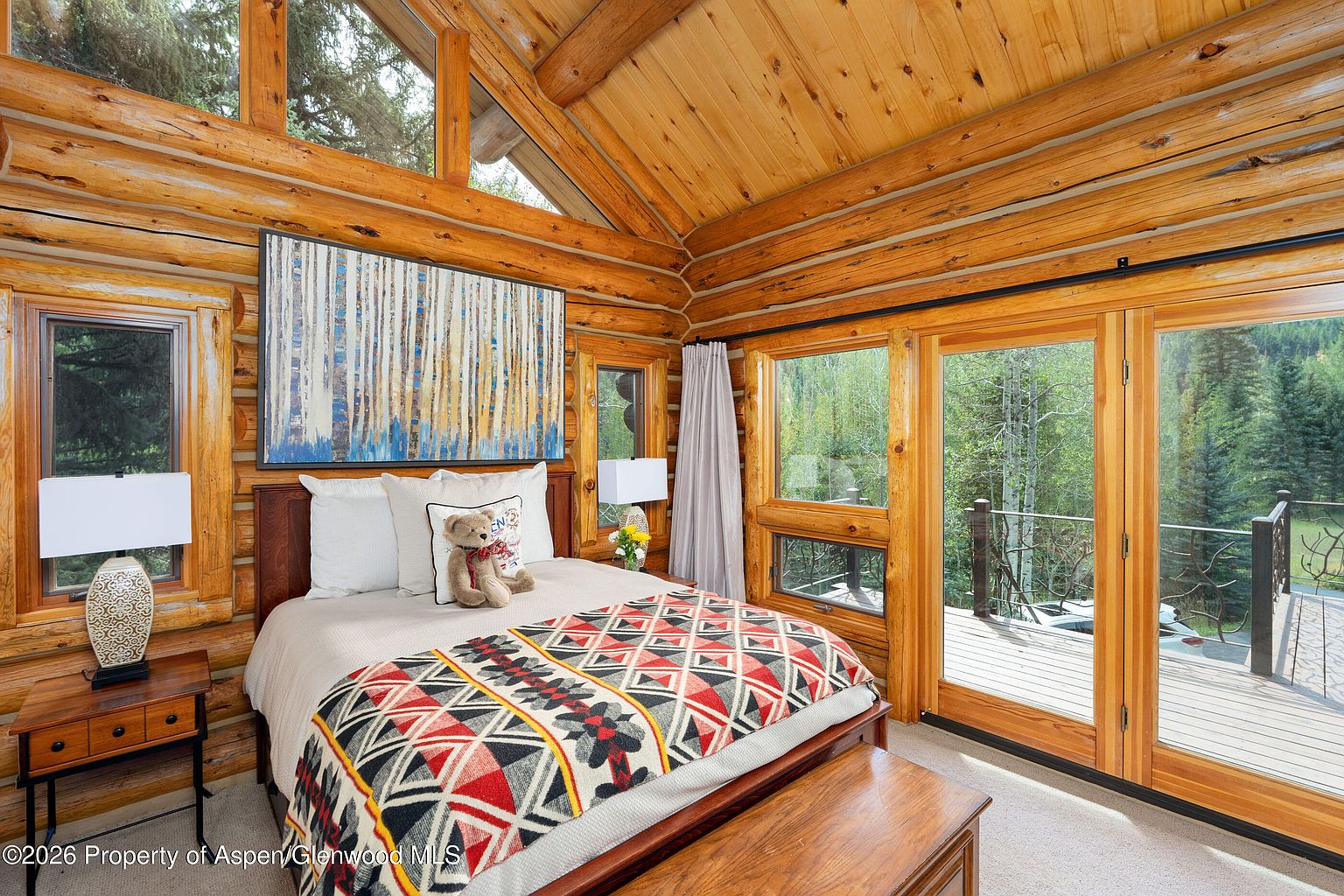 This cozy bedroom features rustic log walls and a vaulted ceiling, creating a warm, cabin-like atmosphere. A bed with a vibrant, patterned quilt is centered in the room, flanked by wooden nightstands and lamps, while large glass doors provide direct access to an outdoor deck with forest views. A piece of abstract art depicting a birch forest hangs above the headboard, enhancing the natural, serene aesthetic of the space.