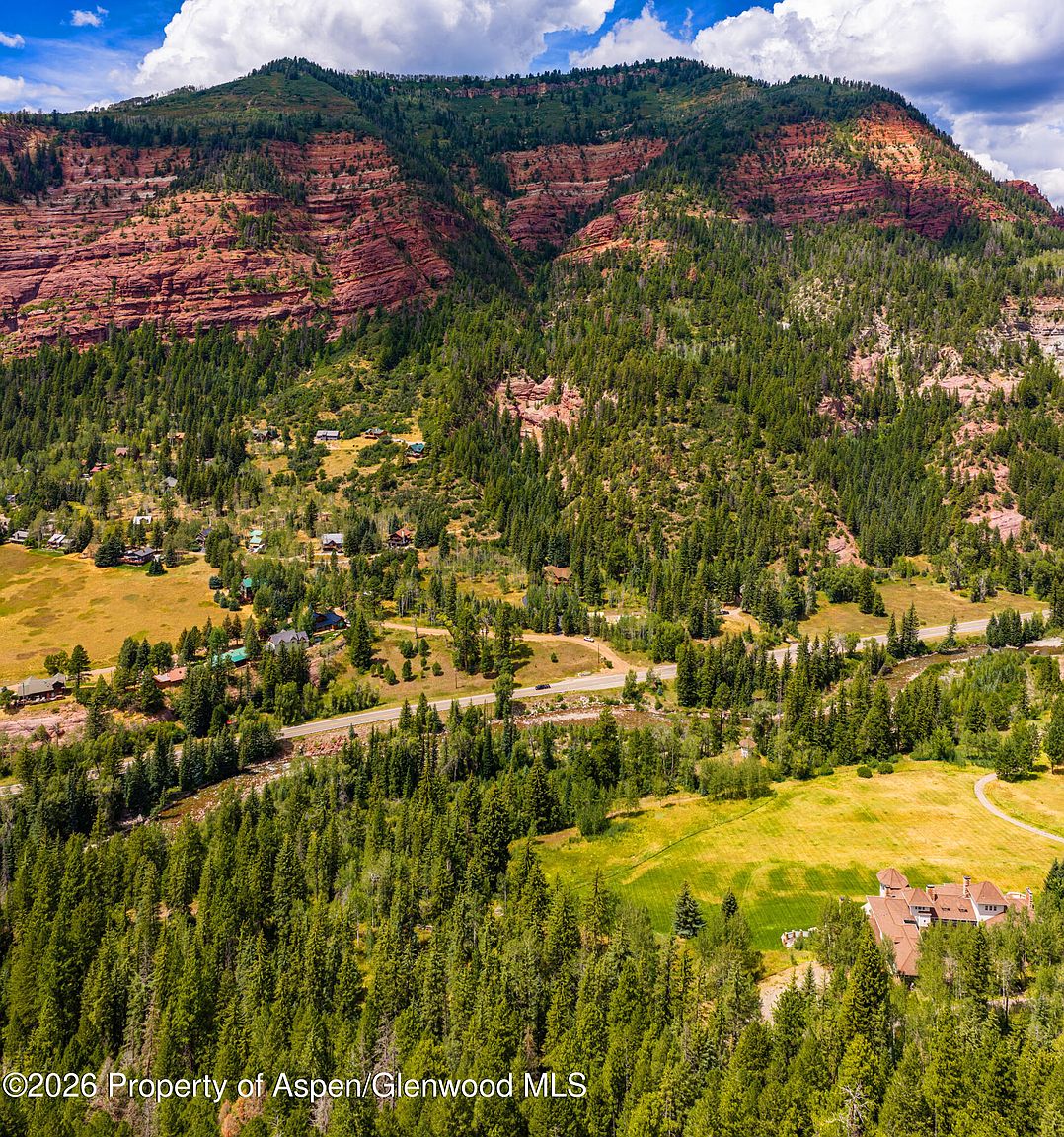 This high-altitude aerial shot captures a sprawling mountain landscape featuring a large, multi-gabled residence nestled among dense evergreen forests and open meadows. A winding road cuts through the valley floor, separating the residential area from the dramatic, rugged red rock cliffs that dominate the background. The perspective provides a grand sense of scale, highlighting the property's secluded location within a vast, natural wilderness.