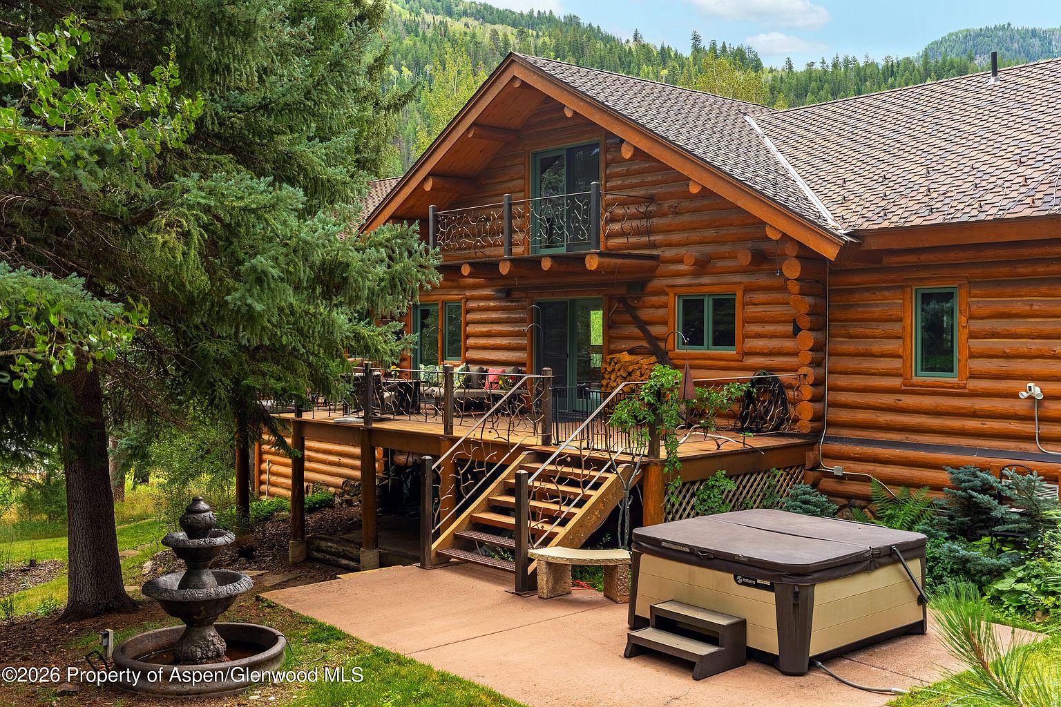 This rustic log cabin features a spacious wooden deck with decorative metal railings and a set of stairs leading down to a concrete patio. The patio area includes a hot tub and a tiered stone fountain, all set against a backdrop of lush green trees and a forested hillside. The home's warm wood exterior and large windows create an inviting, secluded mountain retreat atmosphere.