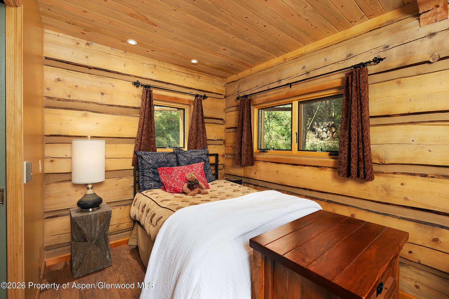 This cozy bedroom features rustic log walls and a matching wood-paneled ceiling, creating a warm, cabin-like atmosphere. The room is furnished with a bed dressed in patterned bedding, a white throw blanket, and a stuffed teddy bear, complemented by a bedside table with a lamp and a wooden chest at the foot of the bed. Two windows with dark patterned curtains allow natural light to filter in, providing a view of the wooded exterior.