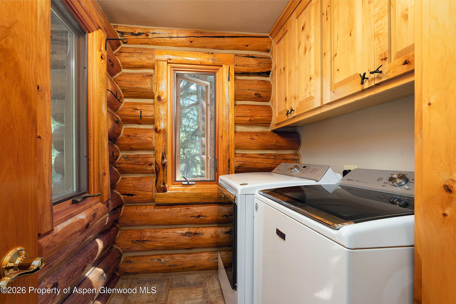 This rustic laundry room features distinctive log walls and warm wood cabinetry, creating a cozy, cabin-like atmosphere. The space is equipped with a white washer and dryer set positioned beneath upper cabinets, with a window providing natural light and a view of the outdoors. The room is finished with neutral-toned floor tiles, emphasizing a functional yet charming aesthetic.