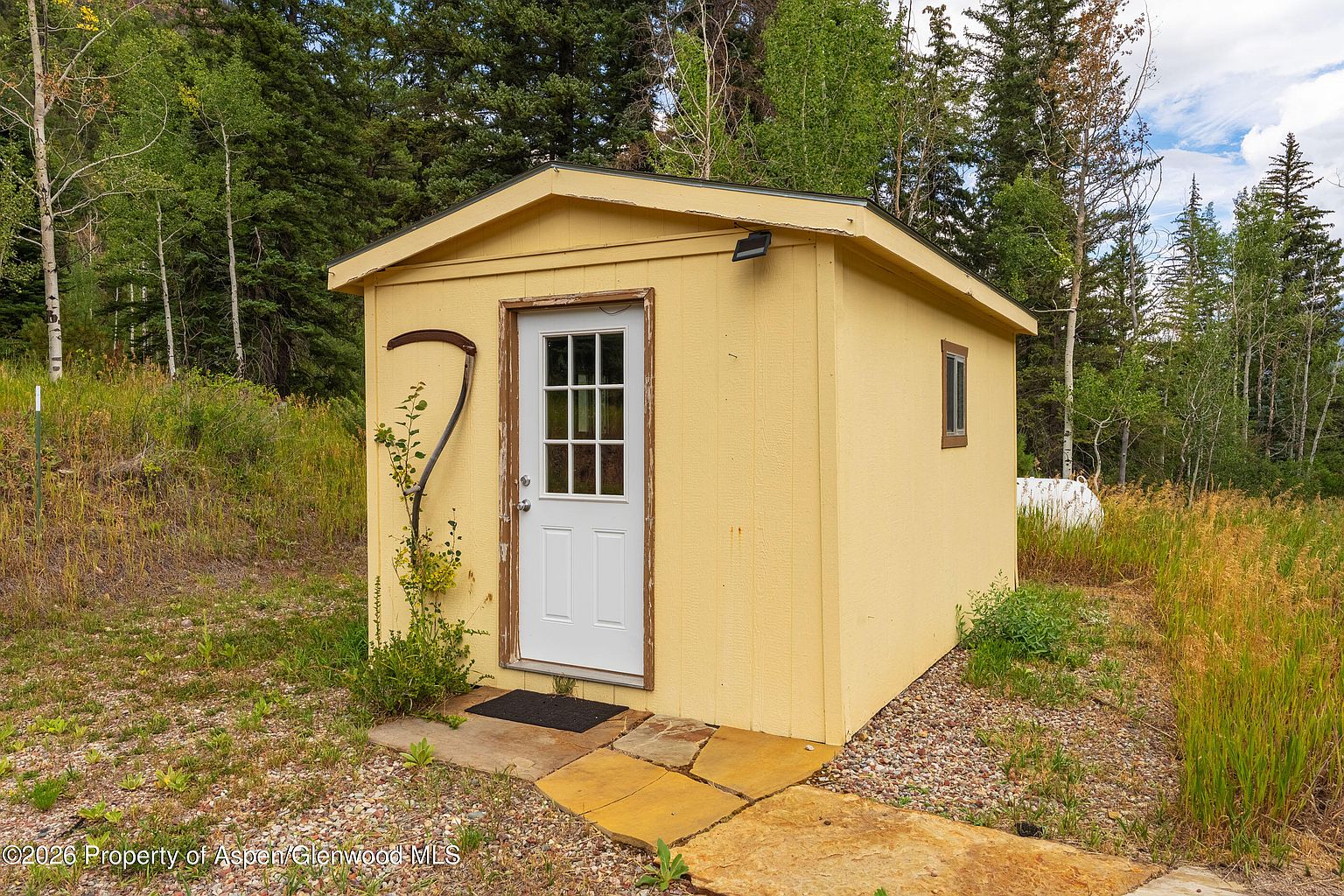 This image features a small, yellow-painted storage shed situated in a natural, wooded setting. The structure has a simple gabled roof, a white door with a window pane, and a single side window, with a rustic scythe mounted on the exterior wall. A stone path leads to the entrance, which is surrounded by tall grasses and dense forest trees in the background.