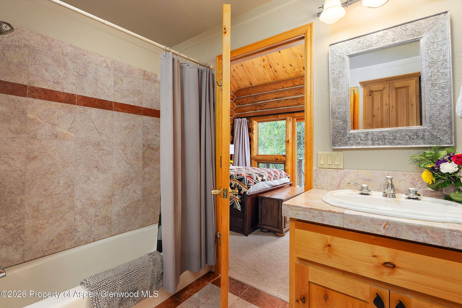 This bathroom features a vanity with a light wood cabinet and a patterned stone countertop, paired with a decorative silver-framed mirror. A shower with a neutral-toned tiled surround and a gray curtain is positioned next to an open doorway that reveals a rustic bedroom with log walls and a view of the outdoors. The perspective captures the transition between the functional bathroom space and the cozy, cabin-style sleeping area.
