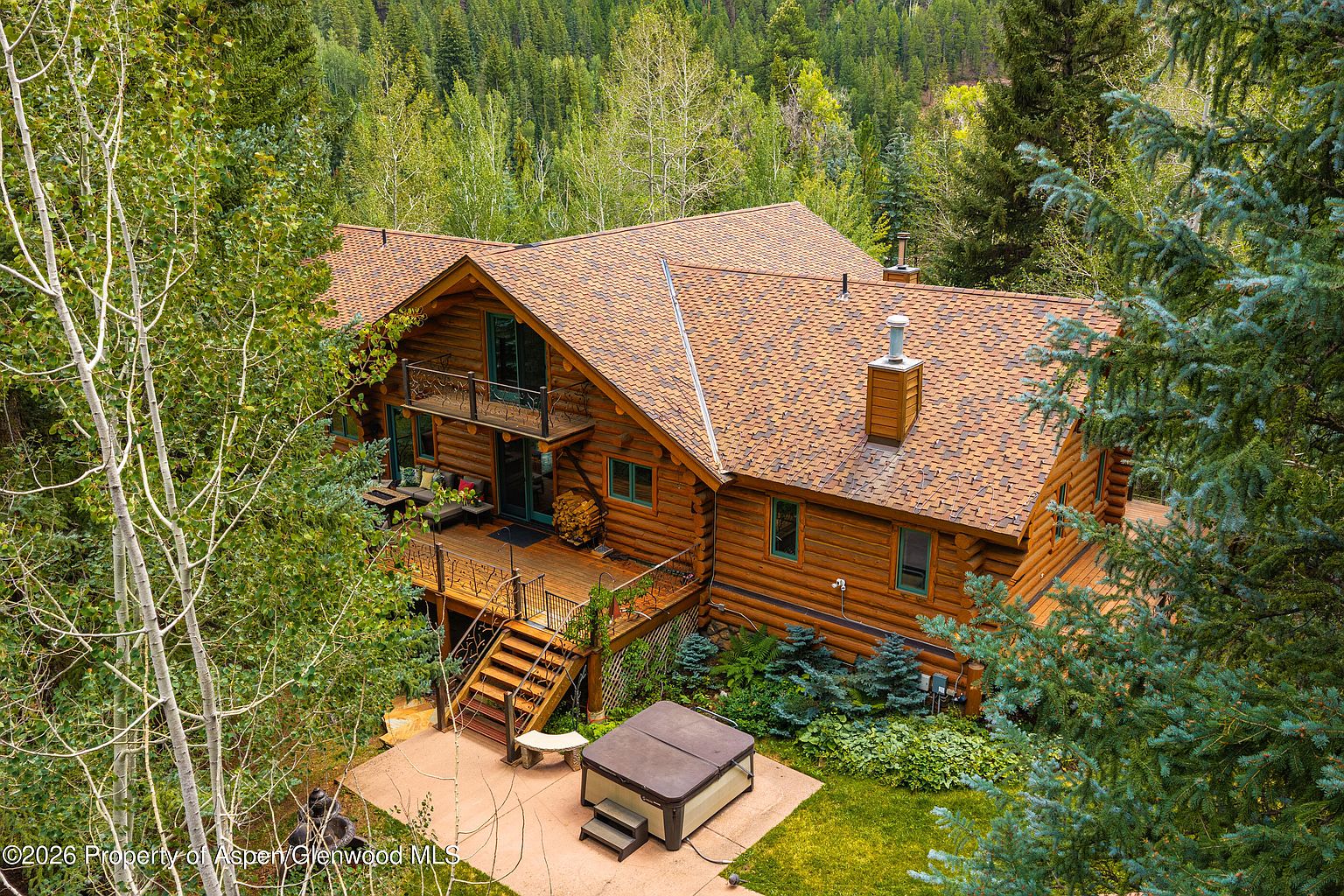 This elevated aerial perspective captures a rustic log cabin nestled within a dense, lush forest. The home features a multi-level wooden deck, a balcony, and a stone chimney, with a hot tub situated on a paved patio area below. The surrounding landscape is filled with mature trees, creating a secluded and serene mountain retreat atmosphere.