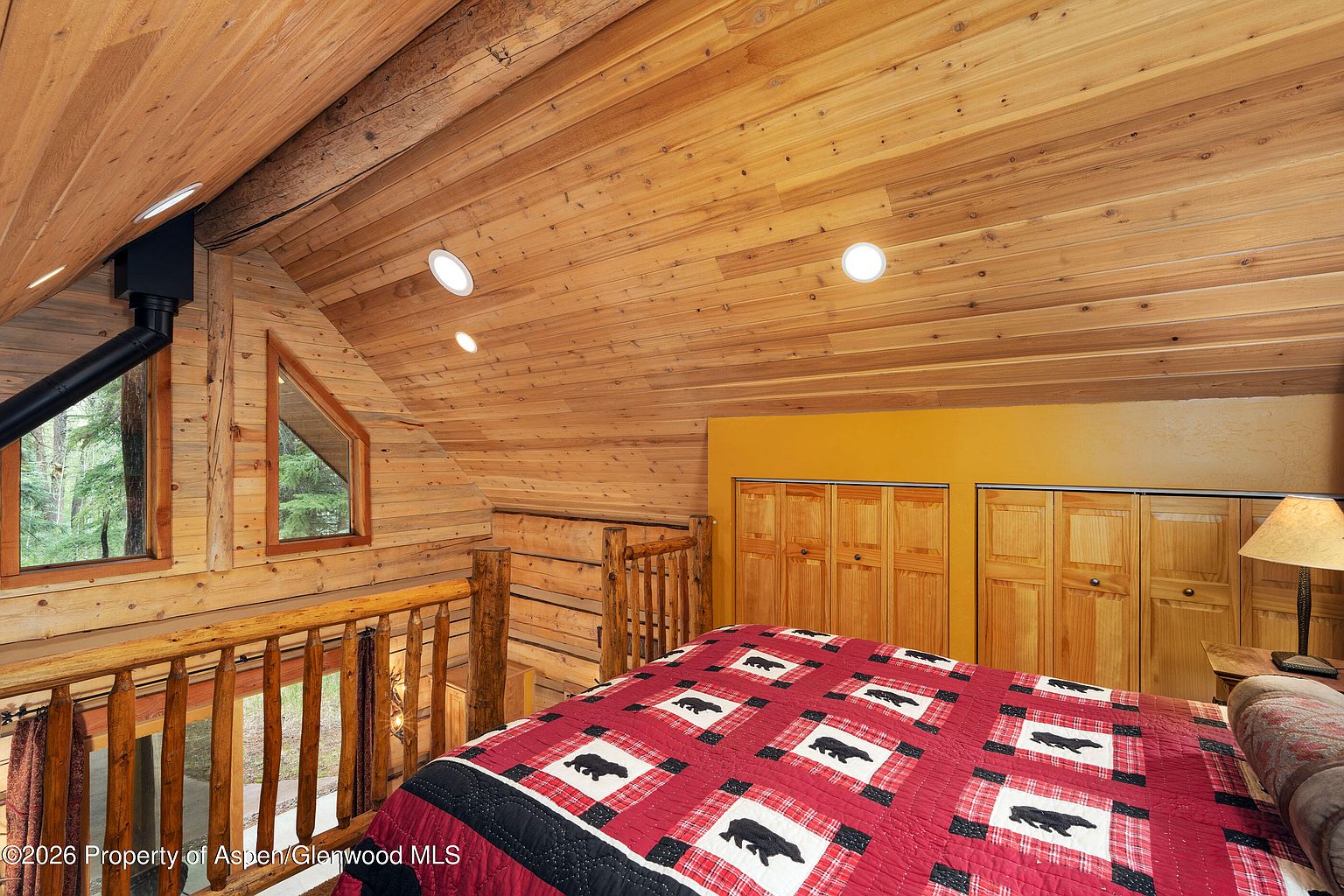 This cozy loft bedroom features a rustic aesthetic with warm wood-paneled walls and a sloped ceiling. A bed with a red and black bear-patterned quilt sits prominently in the foreground, while a wooden railing overlooks the lower level of the home. The space is accented by a vibrant yellow wall section, built-in wooden closet doors, and a bedside lamp, creating a comfortable and inviting cabin atmosphere.