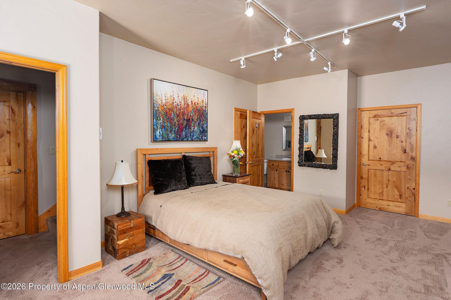 This cozy bedroom features a light wood bed frame with a neutral-toned comforter and dark accent pillows, centered beneath a vibrant abstract painting. The room is illuminated by a modern track lighting system on the ceiling and two bedside lamps, creating a warm and inviting atmosphere. A wooden door leads to an attached bathroom, while a framed mirror hangs on the wall, adding depth to the space.