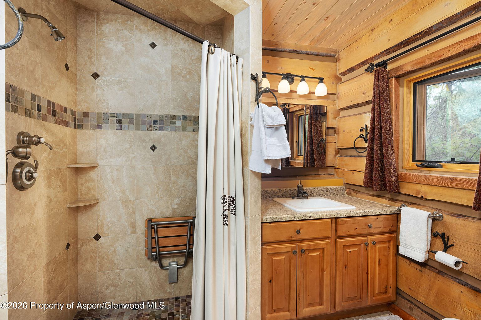 This rustic-style bathroom features log-style walls and a tiled walk-in shower equipped with a fold-down wooden bench. A vanity with a stone countertop and a single sink sits adjacent to the shower, complemented by a mirror and a multi-light fixture. The space is accented with warm wood tones and a window that brings in natural light, creating a cozy and functional atmosphere.