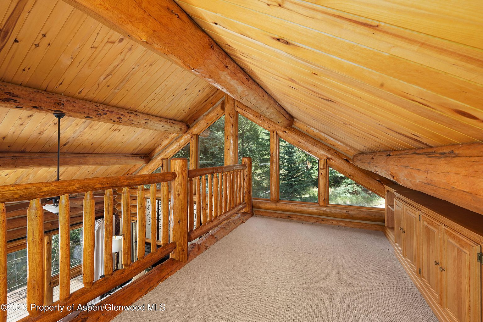This rustic loft area features exposed log beams and light wood paneling on the vaulted ceiling, creating a warm and cozy atmosphere. A wooden railing overlooks the level below, while large windows provide a view of the surrounding trees. The space is finished with neutral carpeting and built-in wooden cabinetry along the side wall.