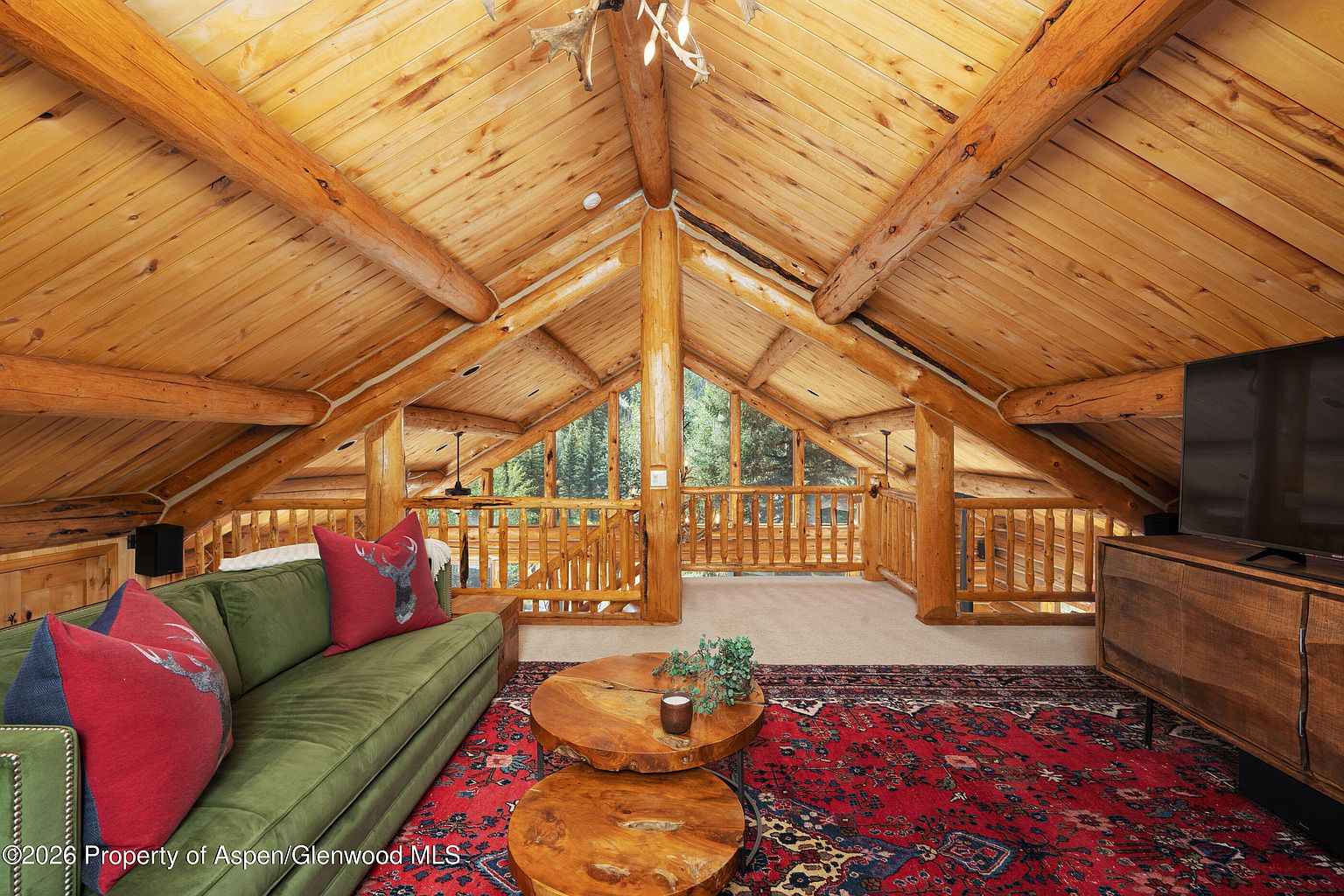 This cozy loft-style living area features a vaulted ceiling with exposed log beams and a rustic aesthetic. A green sofa with red accent pillows sits atop a large, intricate red patterned rug, complemented by two round wooden coffee tables. The space opens to a wooden railing overlooking the level below, with large windows providing a view of the surrounding forest.